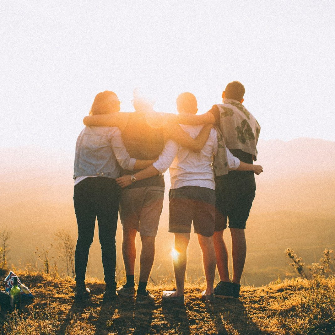 four person hands wrap around shoulders while looking at sunset